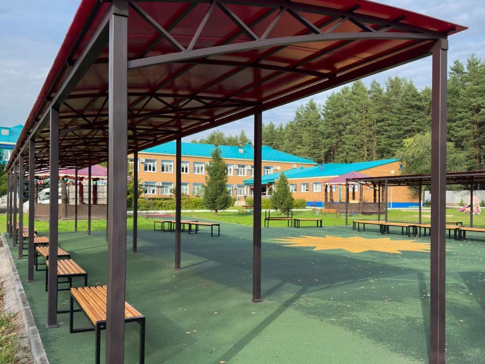Длинный навес с красной арочной кровлей из поликарбоната над скамейками на школьном дворе с мягким резиновым покрытием
